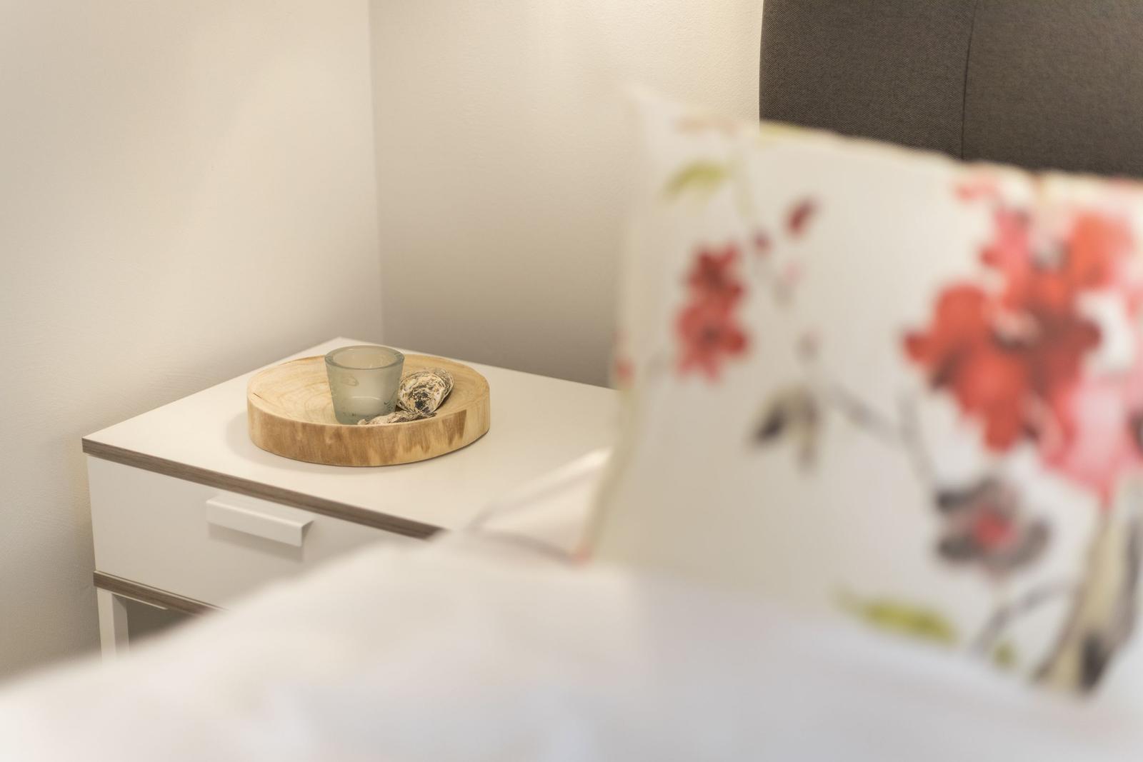 Nachttisch mit Holztablett, Glas und Muscheln. Kissen mit Blumenmuster im Vordergrund.