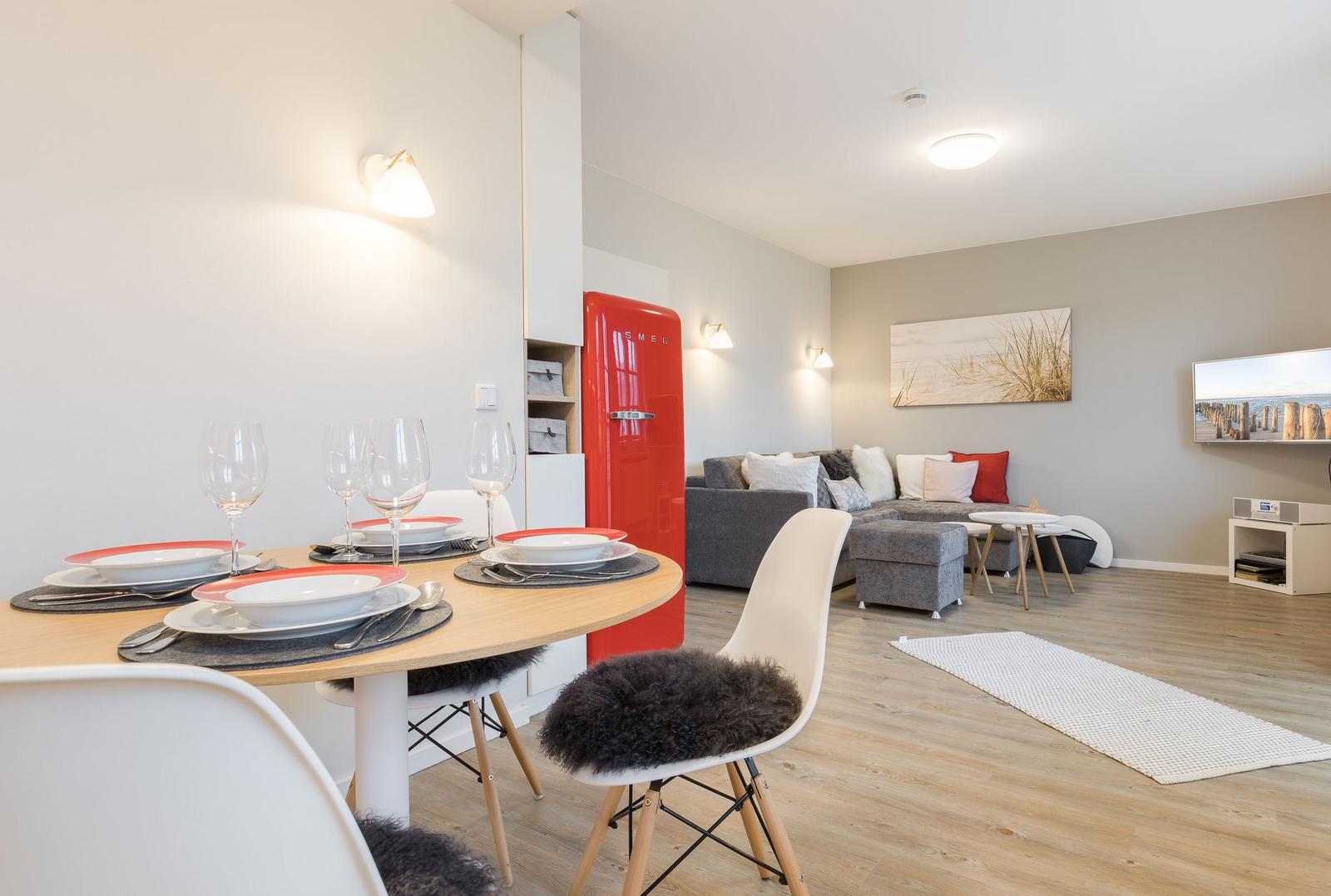 Essbereich mit runden Tisch und Stühlen. Im Hintergrund Wohnbereich mit Sofa und rotem Kühlschrank.
