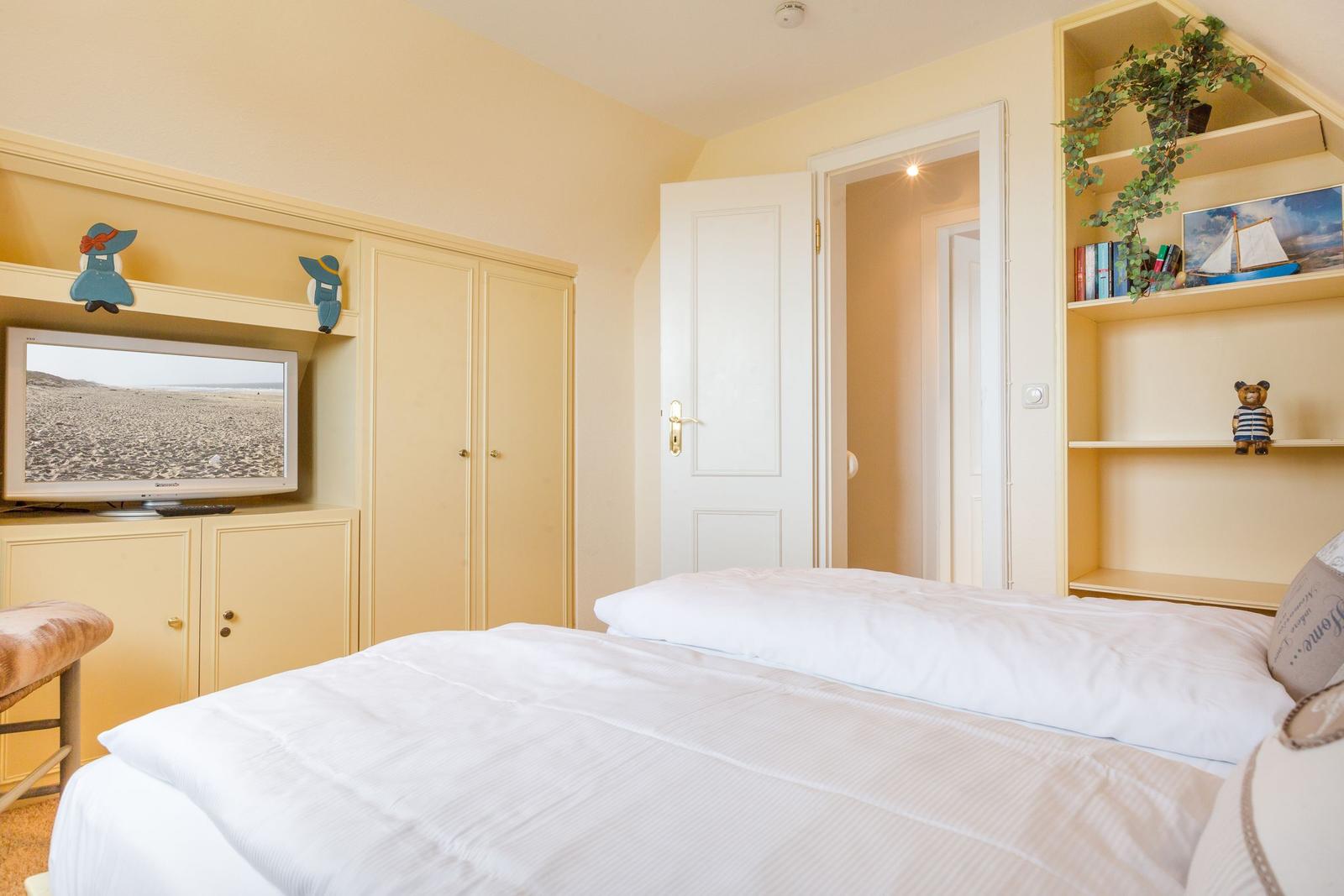 Schlafzimmer mit Bett, TV und Regal. Tür führt in angrenzenden Raum.