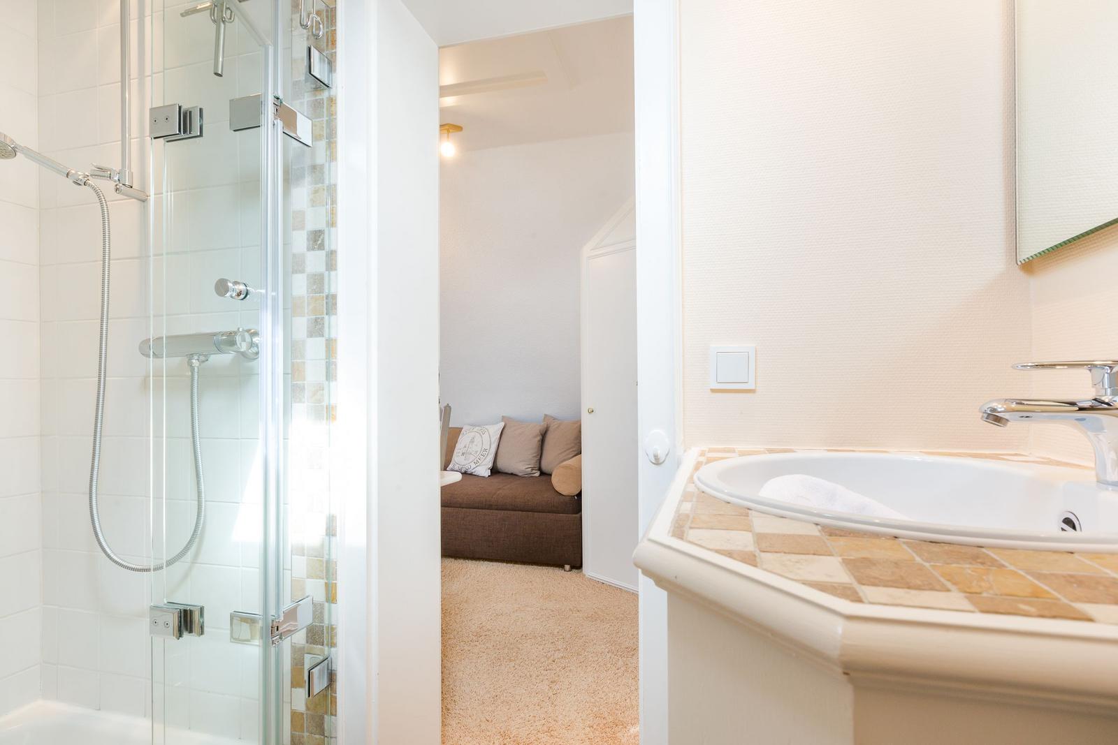 Bad mit Dusche und Waschbecken. Im Hintergrund ist ein Wohnbereich mit Sofa.