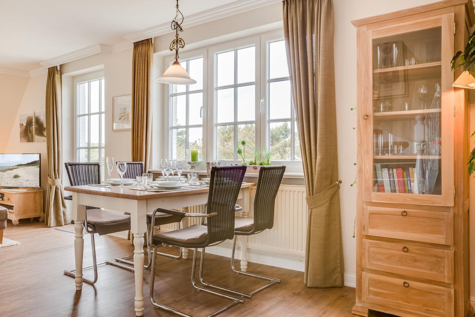 Esszimmer mit Tisch, Stühlen und Glasvitrine. Fenster mit Vorhängen und Blick nach draußen.