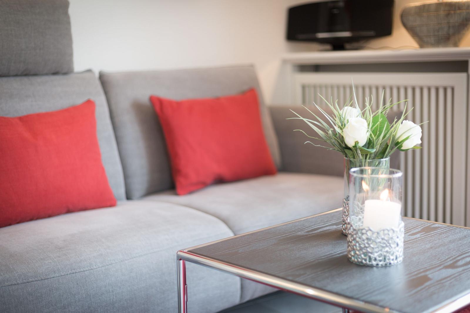 A gray sofa with red cushions and a table with a candle and flowers.