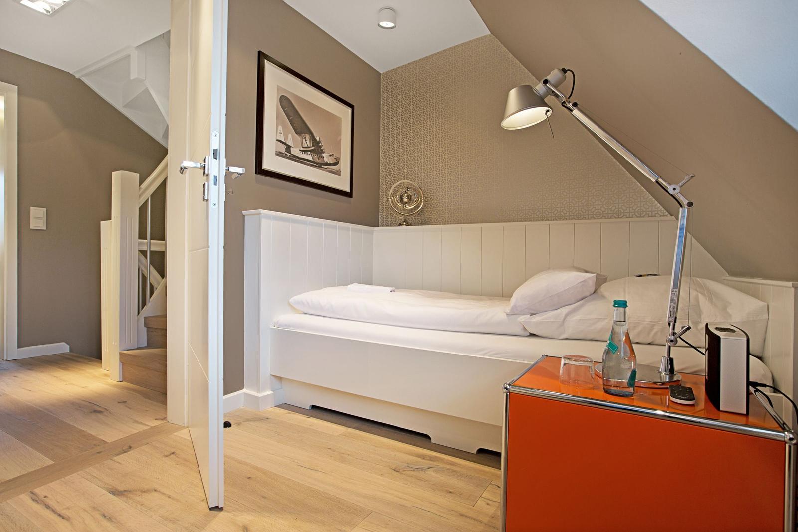 Schlafzimmer mit weißem Bett, orangefarbenem Nachttisch und Stehlampe unter schrägem Dach.