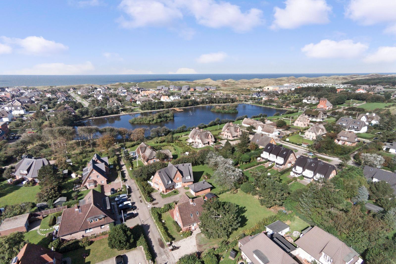 Luftbild von Häusern mit Grünflächen, einem See und dem Meer im Hintergrund.