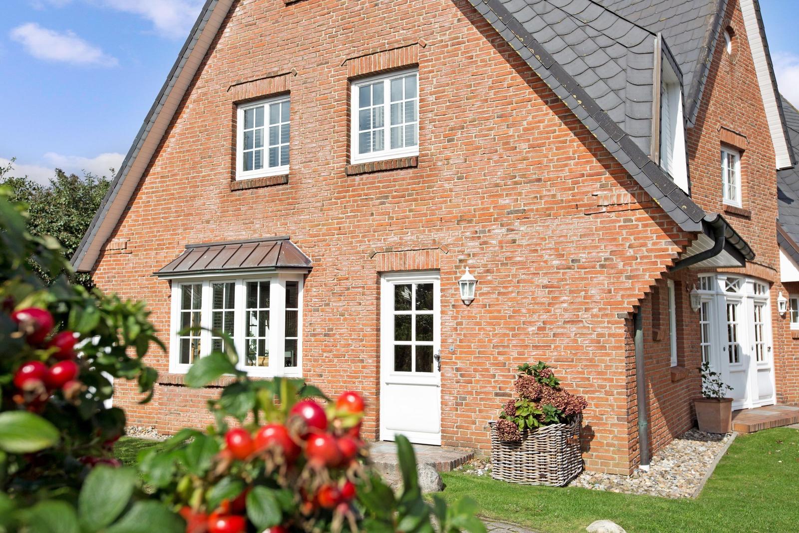 Rotes Backsteinhaus mit weißen Fenstern und Tür. Vorgarten mit Blumenkorb und Beerensträuchern.