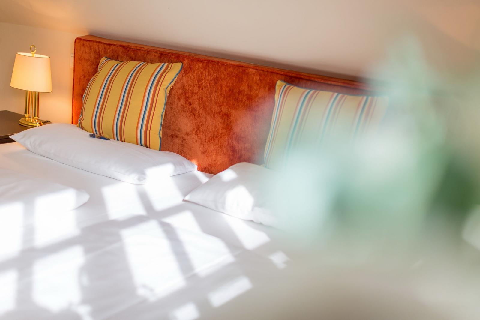 A bed with white sheets and an orange headboard. Two pillows with striped pattern.