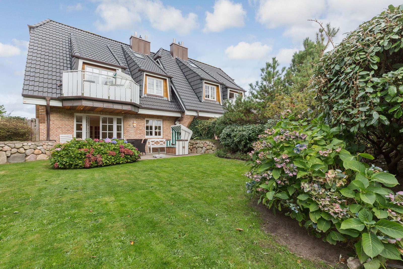 House with balcony, garden, and flower hedges under blue sky.
