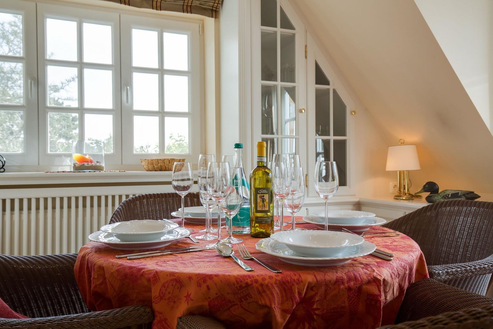 Dining table with tablecloth, glasses, and a wine bottle. Window and cabinet in the background.