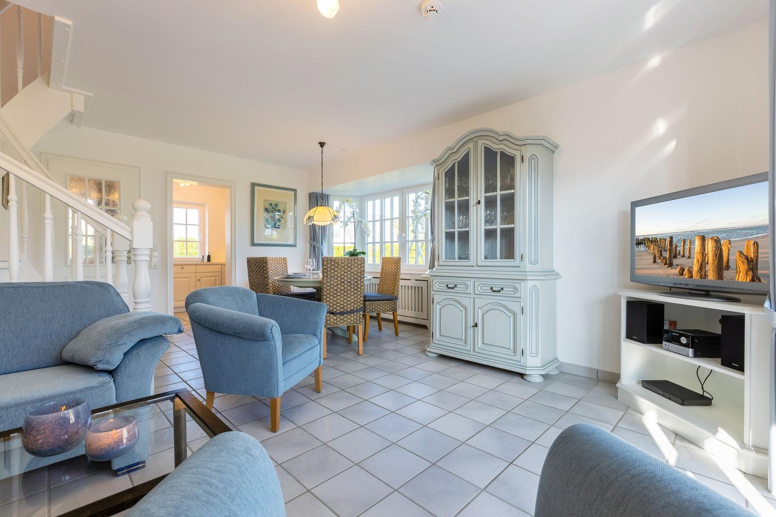 Living and dining area with blue seating, white furniture, and TV. Tile floor and large window.