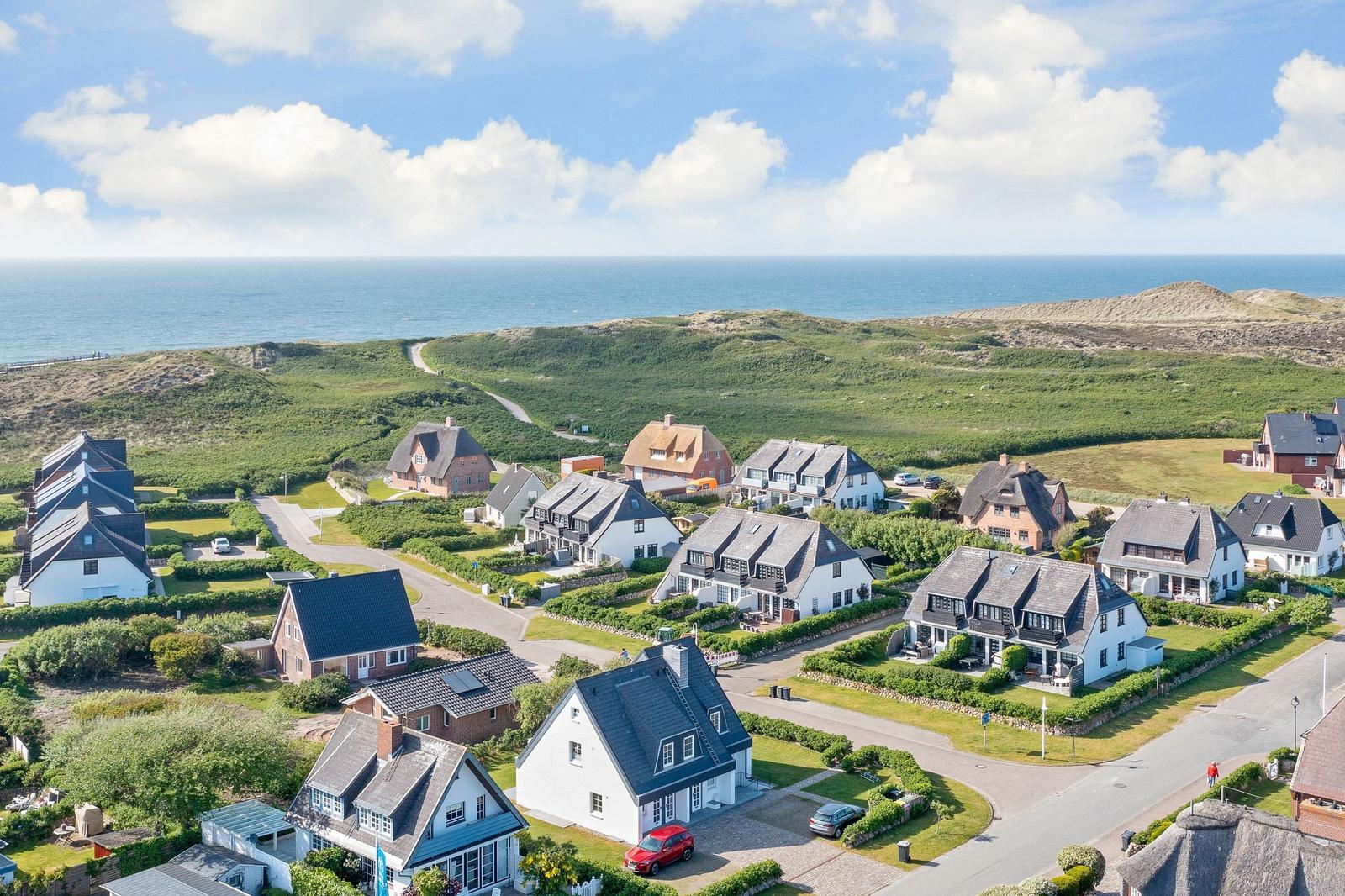 Houses with roofs and gardens near the coast. Sea and green hills in the background.