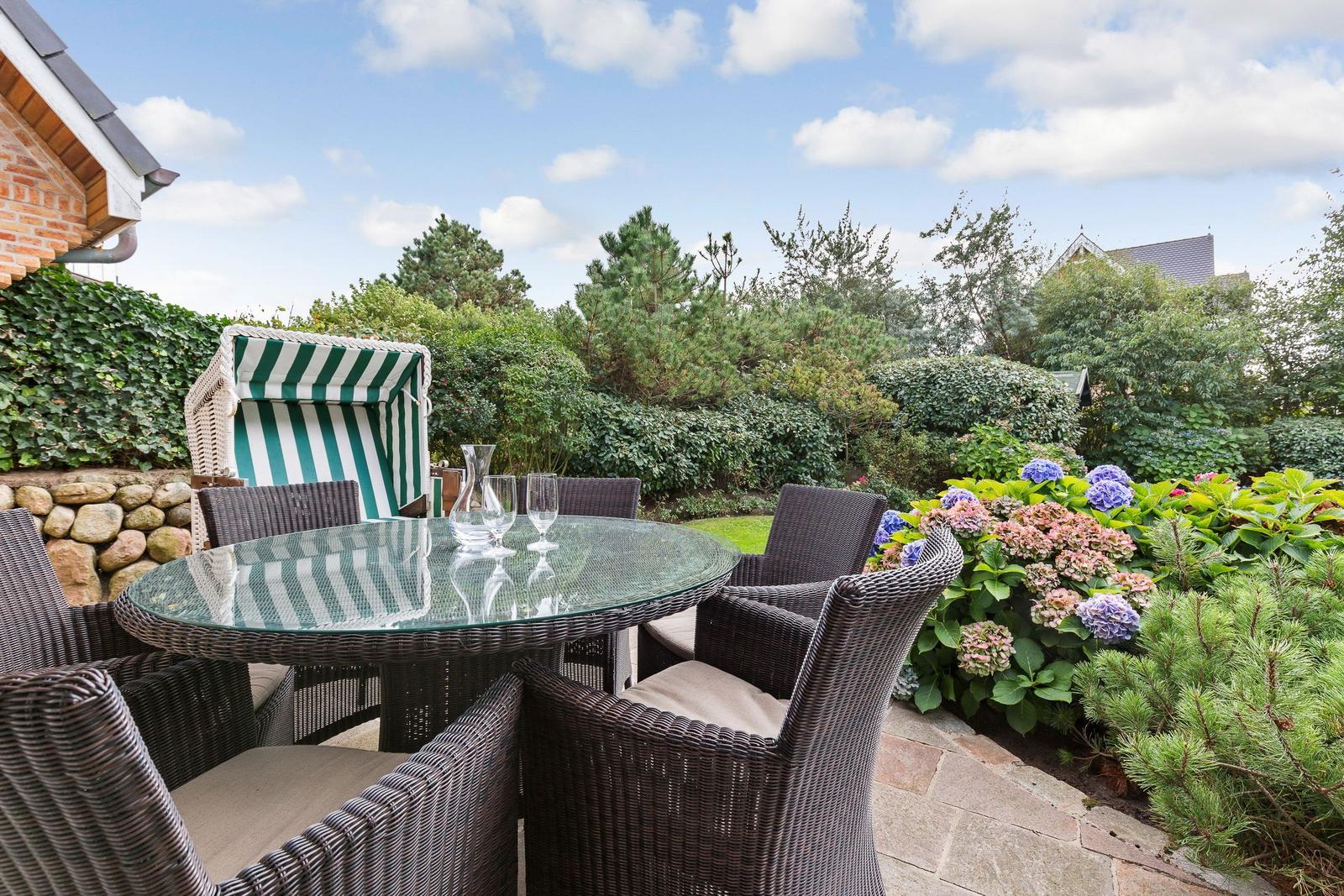 Garden with wicker furniture, glass table, and sunshade. Flowerbeds and trees in the background.