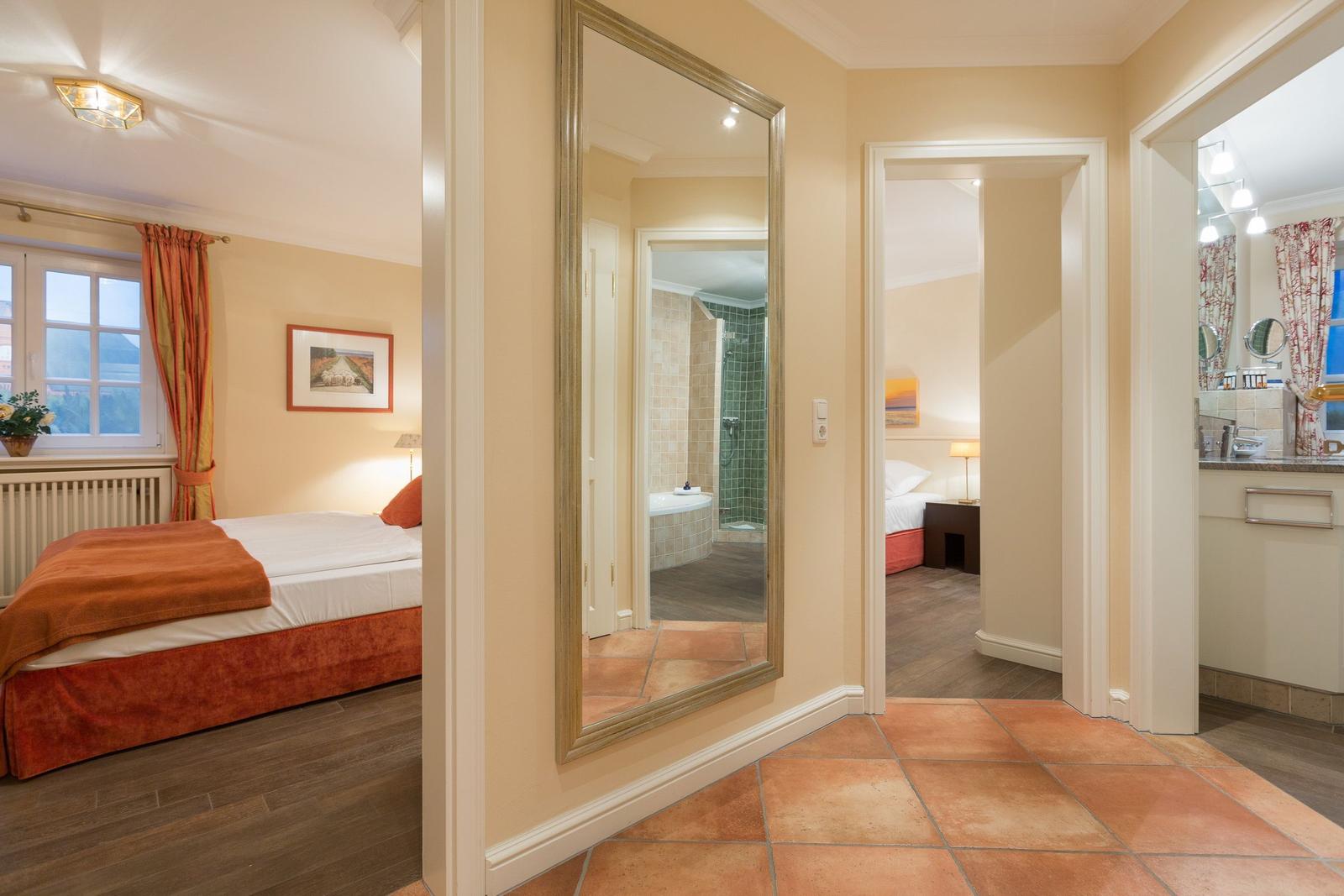 Bedroom with bed and window, bathroom with shower and bathtub, hallway with mirror and terracotta floor.