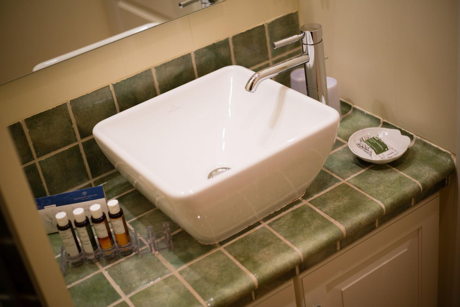 Sink with faucet on green tiled countertop with soap and toiletries.