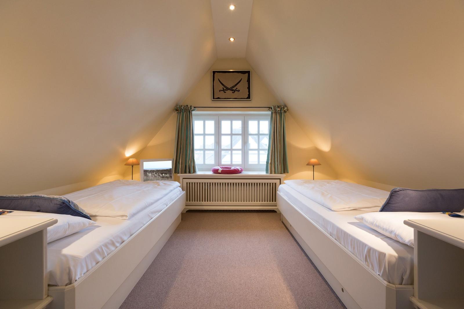 Two beds under sloped ceiling with window and curtains.