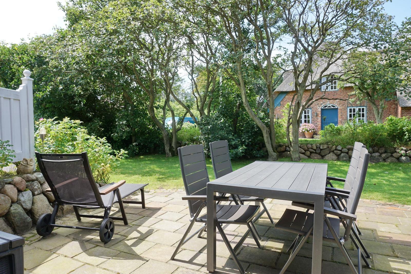 Terrace with table and chairs, surrounded by greenery and a house in the background.