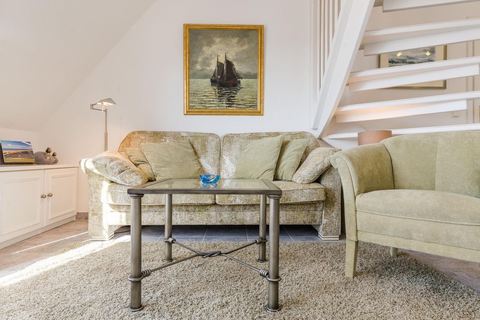 Wohnzimmer mit Sofa, Sessel und Glas-Tisch unter einer Treppe.