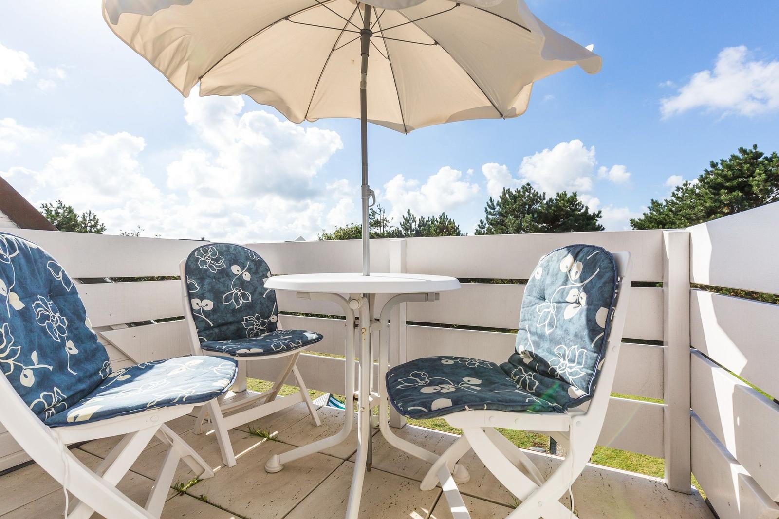 Terrasse mit Tisch, drei Stühlen und Sonnenschirm. Weißer Holzzaun und Bäume im Hintergrund.