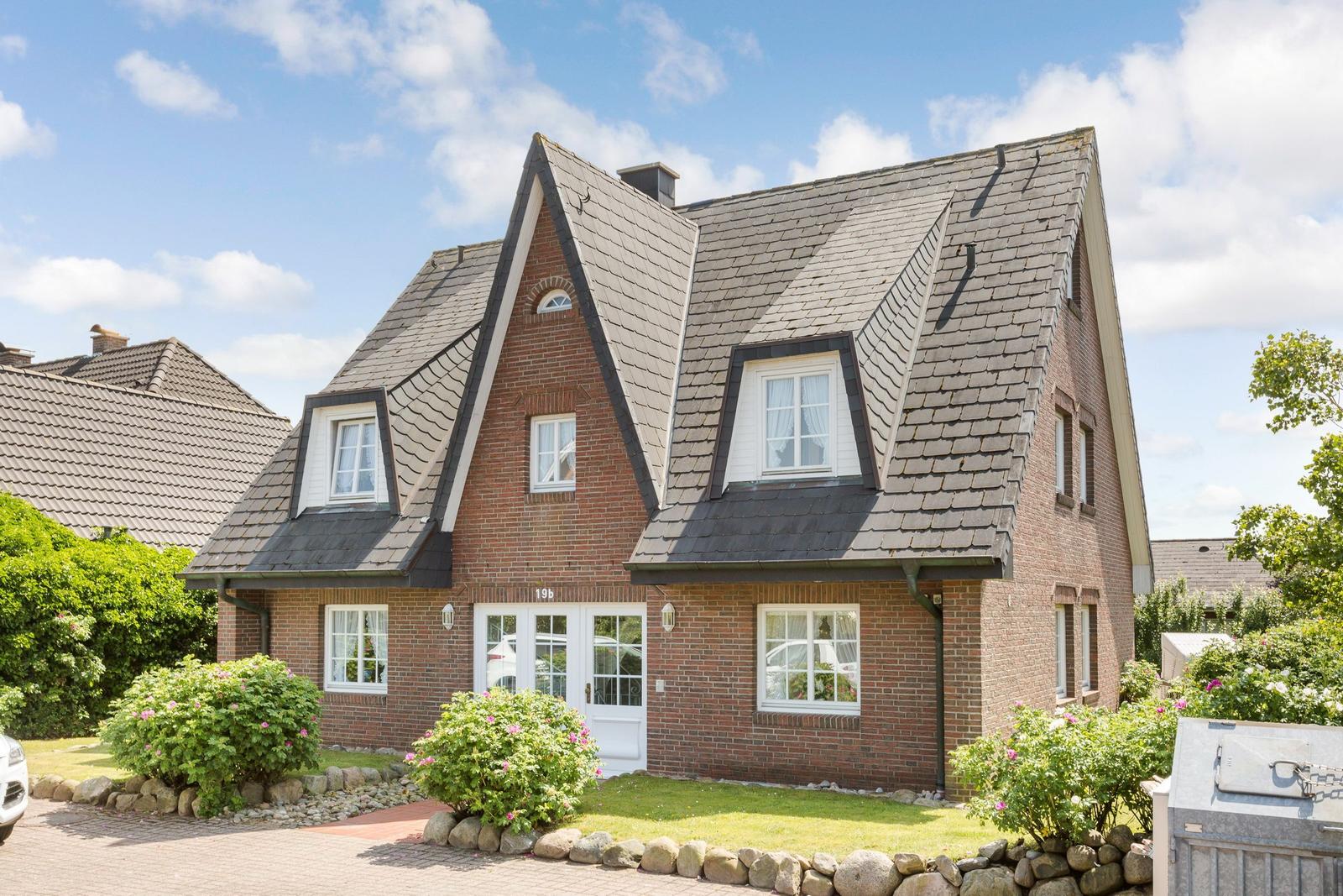 Two-story brick house with dormers and white entrance. Garden with flowers and stone wall.