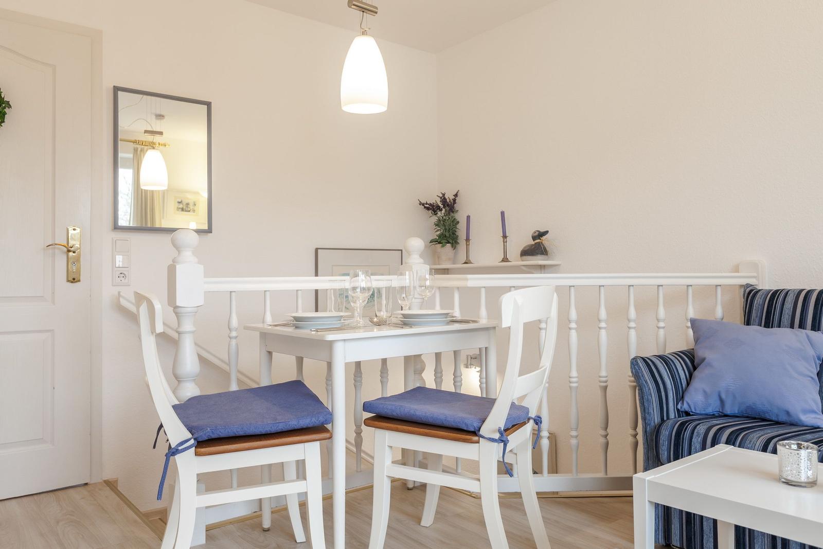 Dining area with white table, two chairs, and blue seat cushions. Background with sofa and railing.