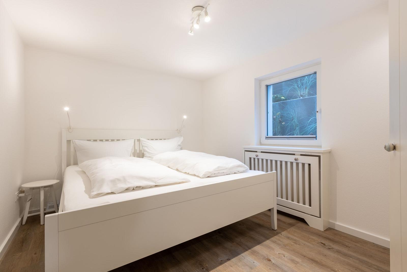 Ein Schlafzimmer mit weißem Bett, Holzboden und Fenster mit Heizkörper.