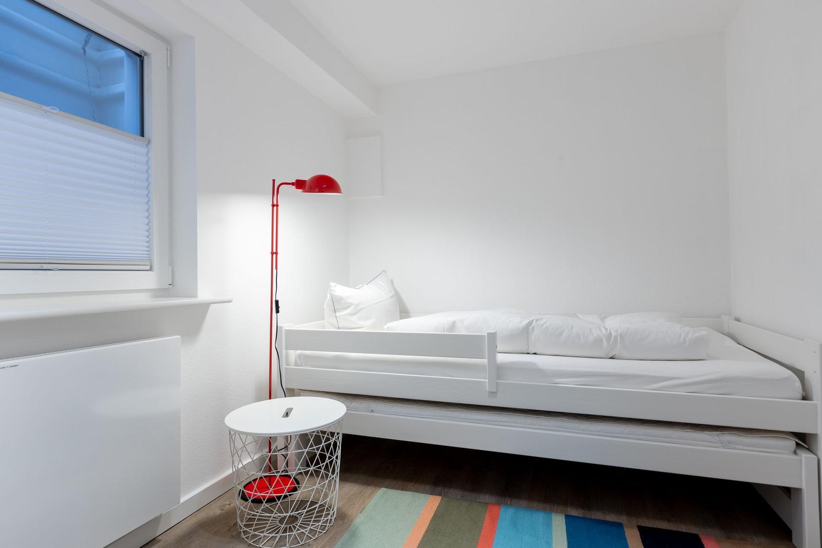 Ein Schlafzimmer mit weißem Bett, roter Stehlampe und buntem Teppich.