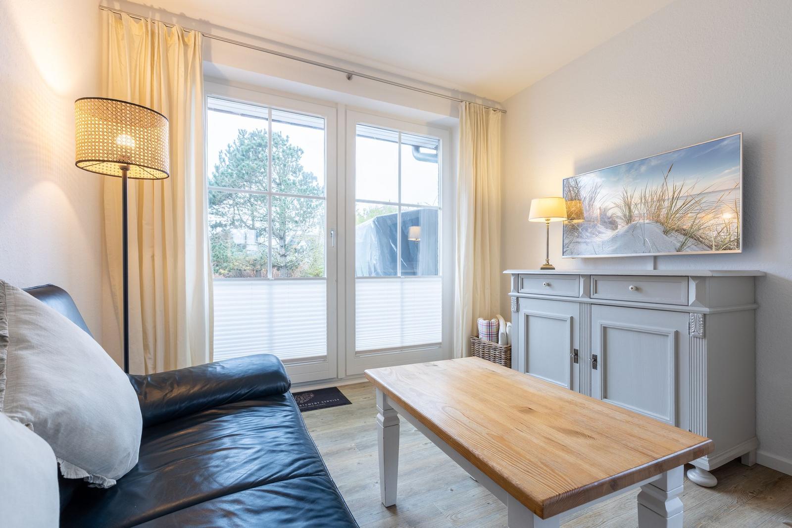 Wohnzimmer mit Sofa, Tisch und TV. Fenster mit Vorhängen und Blick nach draußen.