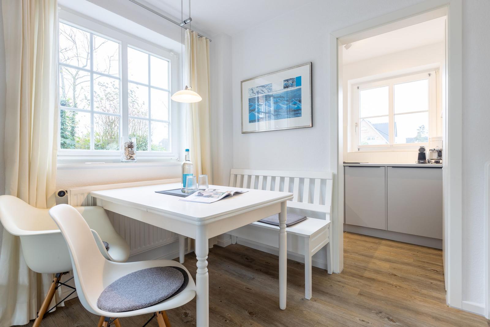 Essbereich mit weißem Tisch, Stuhl und Bank. Küche im Hintergrund. Holzboden und Fenster.