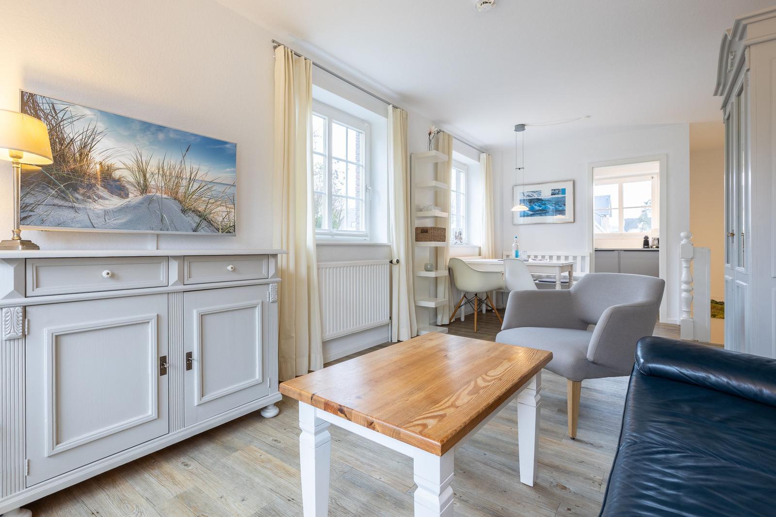 Wohnzimmer mit Holztisch, grauen Sesseln und weißen Schränken. Fenster mit Vorhängen.