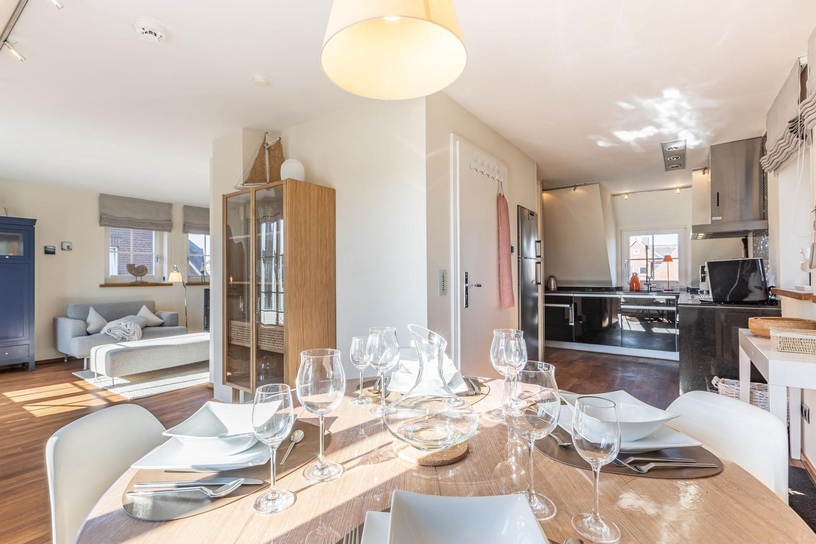 Esszimmer mit Tisch, Stühlen und Gläsern. Küche und Wohnzimmer im Hintergrund.