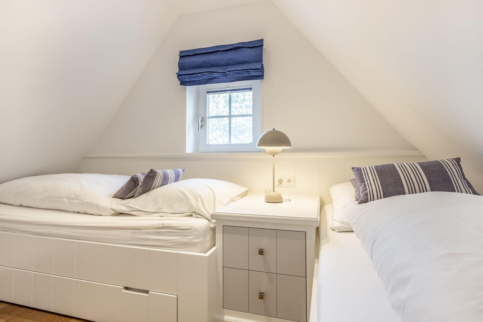 Zwei Betten mit weißen Bettwäsche und gestreiften Kissen. Zwischen den Betten steht ein Nachttisch mit Lampe.