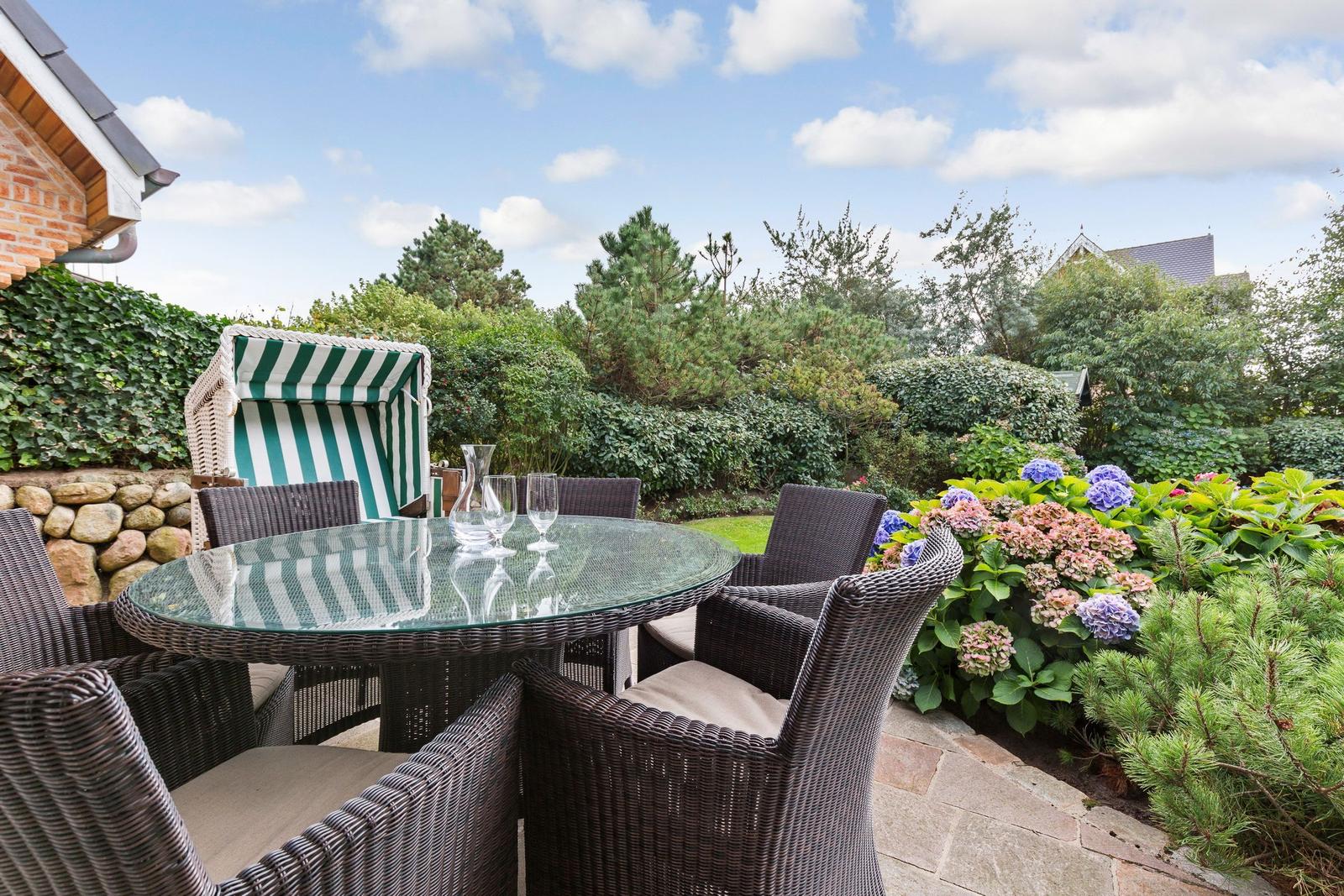Garden with wicker furniture, glass table, and green-and-white lounge chair. Flower beds and trees in the background.