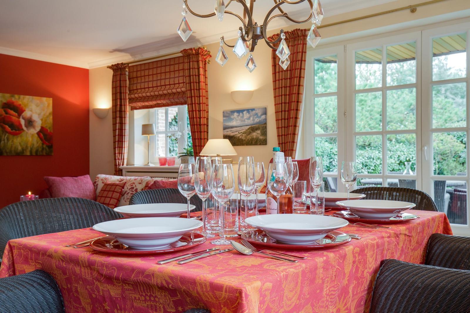 Dining table with tablecloth, glasses, and cutlery. Windows with curtains and chandelier.