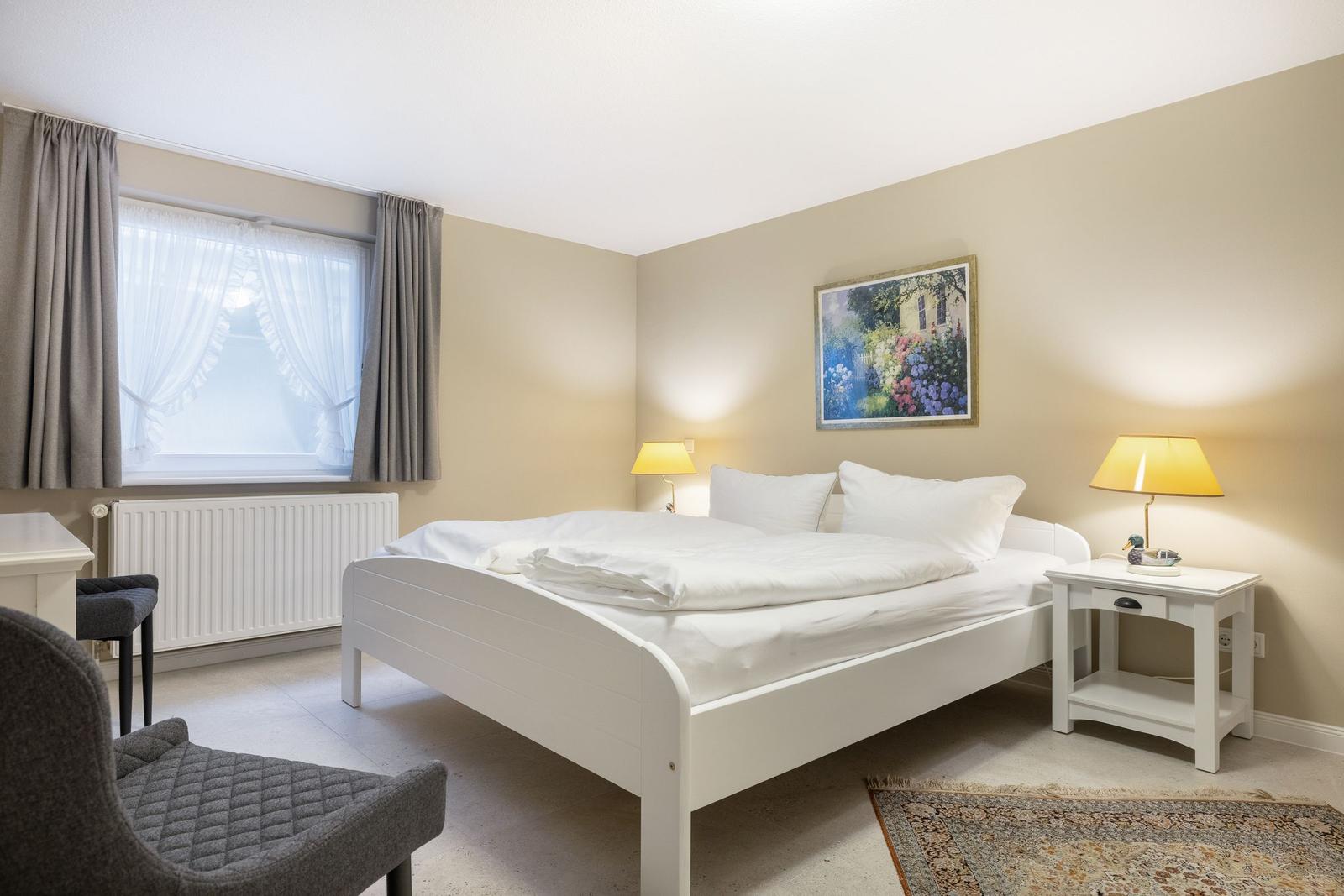 Ein Schlafzimmer mit weißem Bett, Nachttischen und Fenster mit Vorhängen.