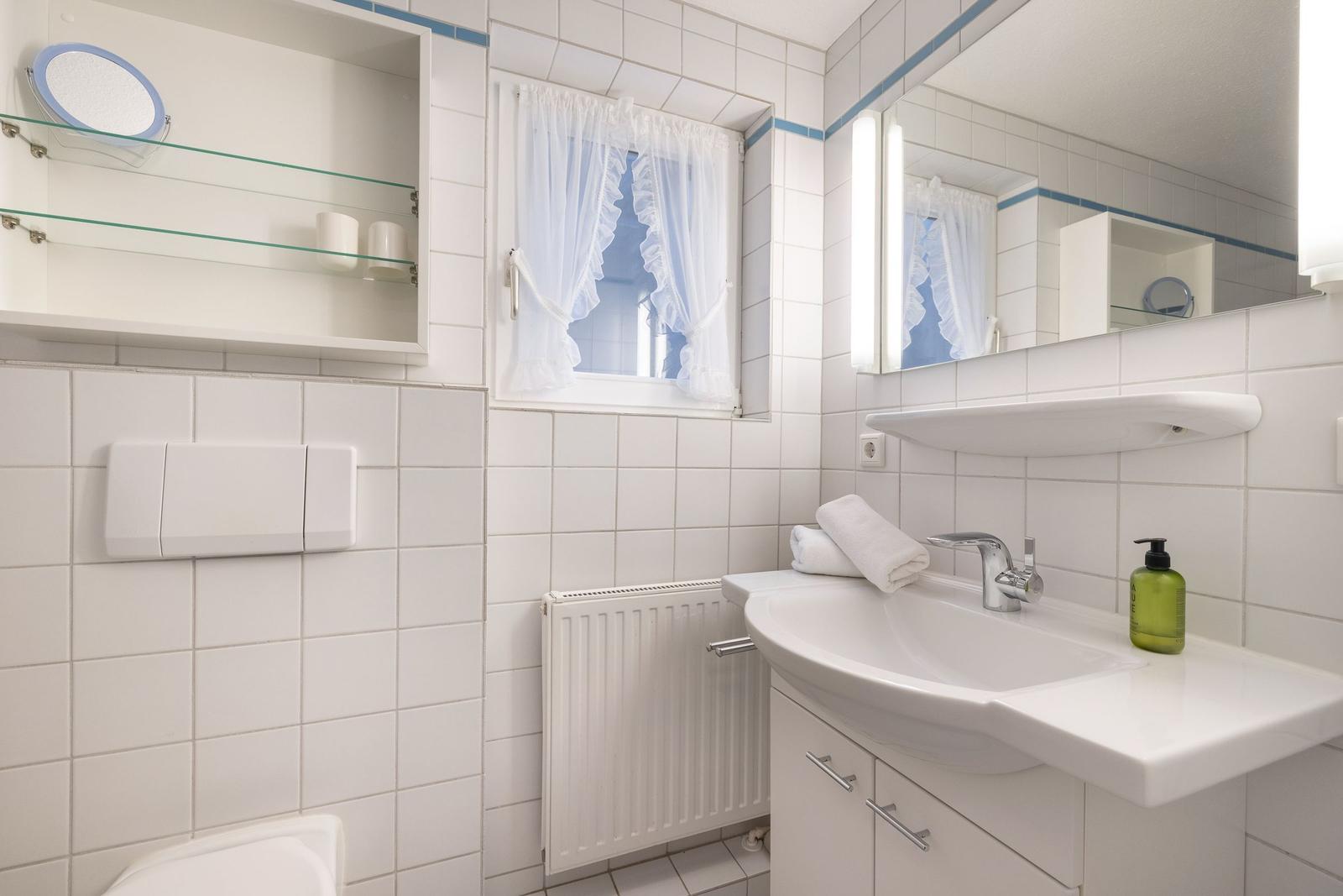 Badezimmer mit Waschbecken, Spiegel und Fenster mit weißen Vorhängen.