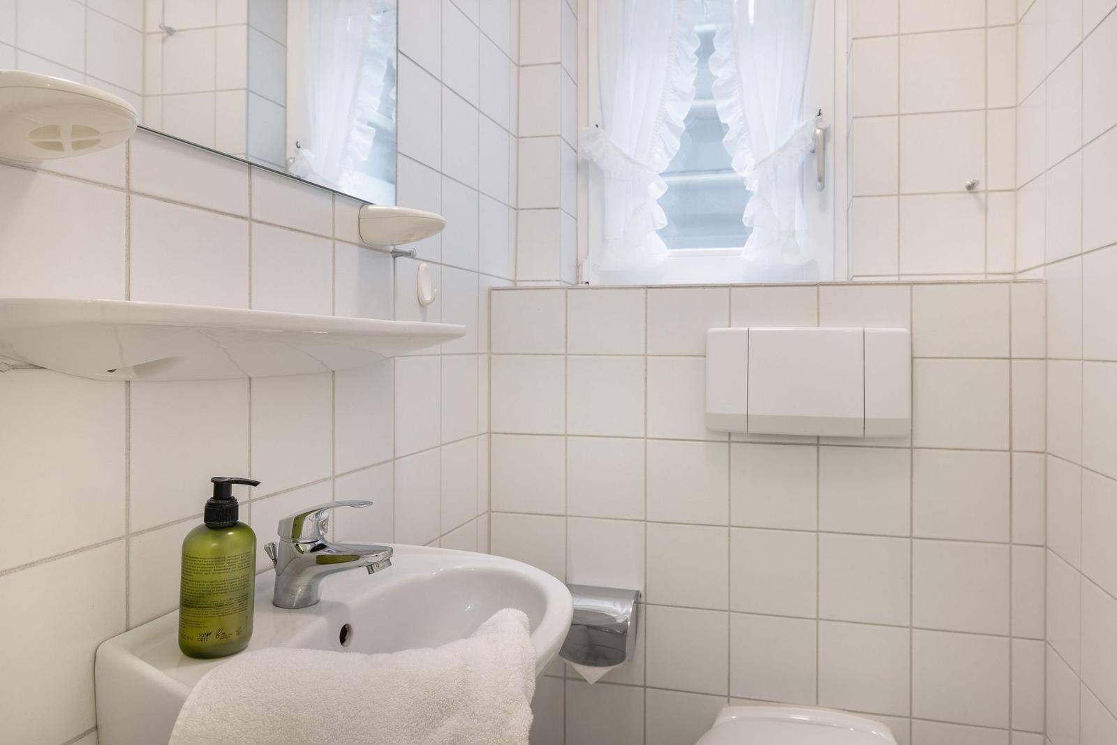Badezimmer mit Waschbecken, Spiegel und Fenster mit weißen Vorhängen.