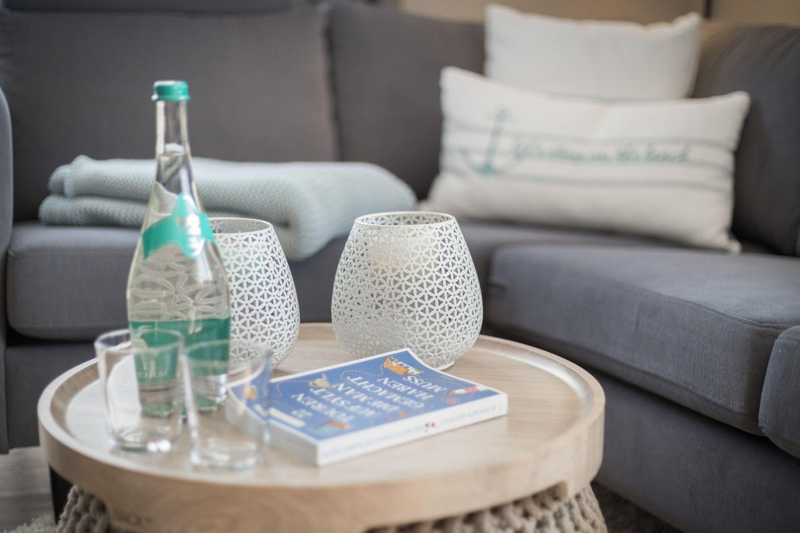 Runder Tisch mit Flasche, Gläsern und Buch vor grauem Sofa.