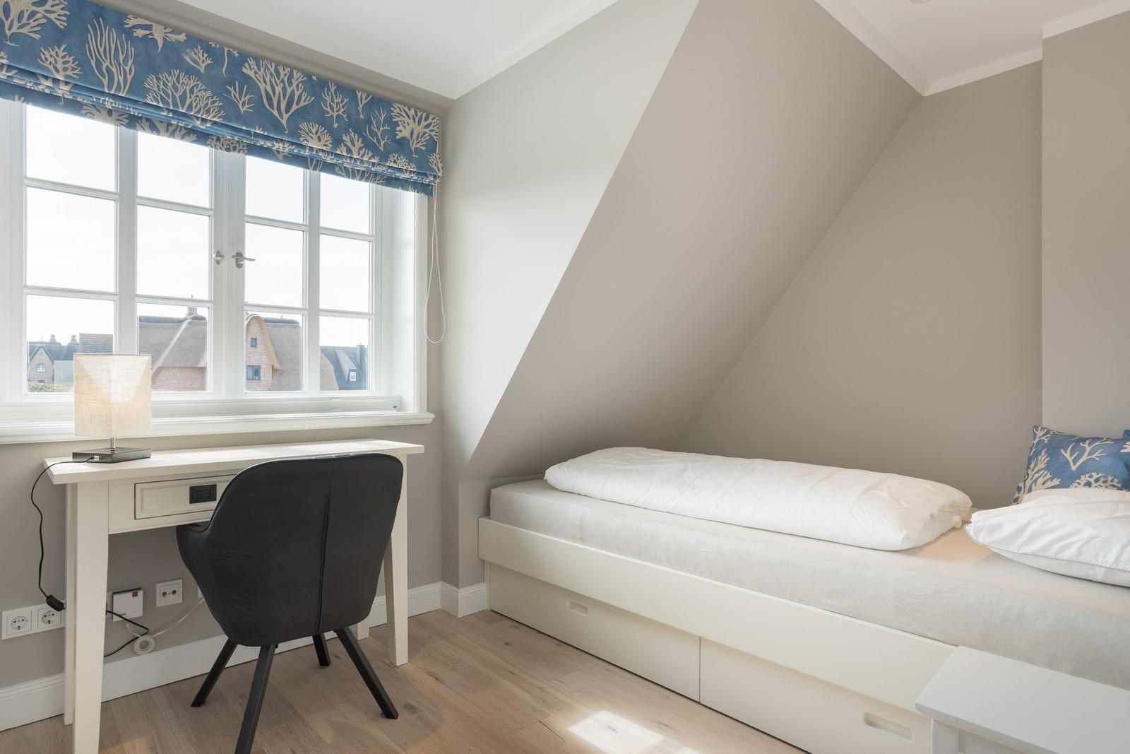 Ein Schlafzimmer mit Dachschräge, weißem Bett und Schreibtisch unter dem Fenster.
