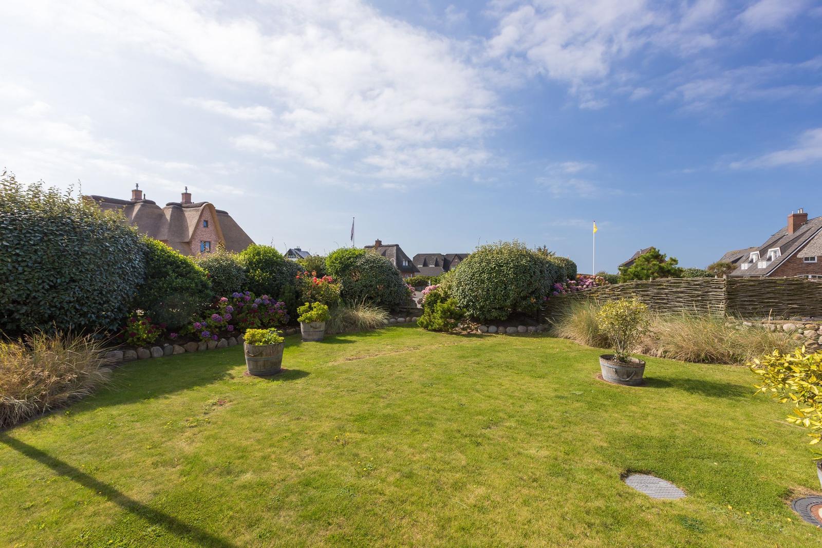 Garten mit Rasen, Blumen und Bäumen. Im Hintergrund sind Häuser zu sehen.