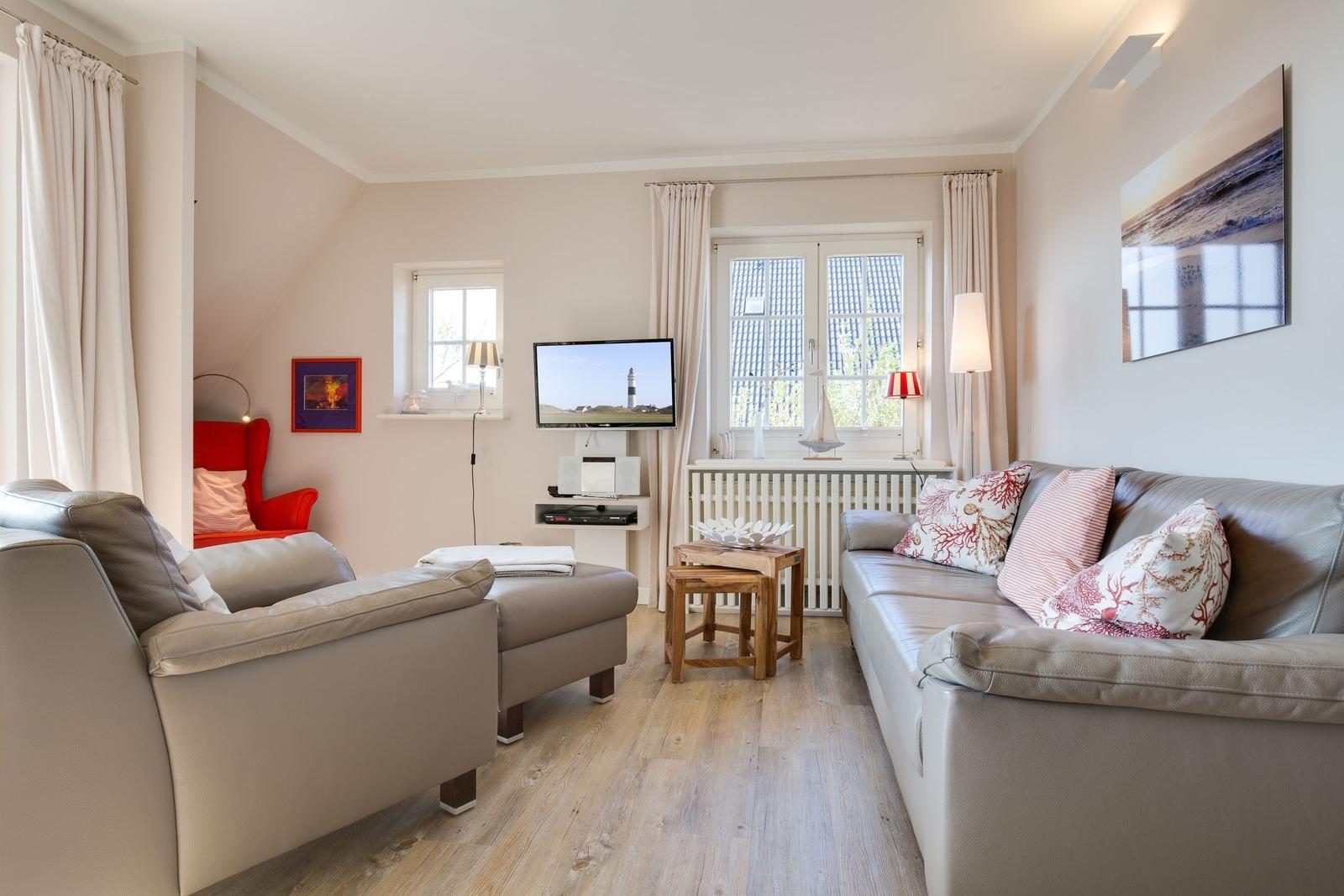 Wohnzimmer mit Sofa, Sessel und Fernseher. Holzboden und Fenster mit Vorhängen.