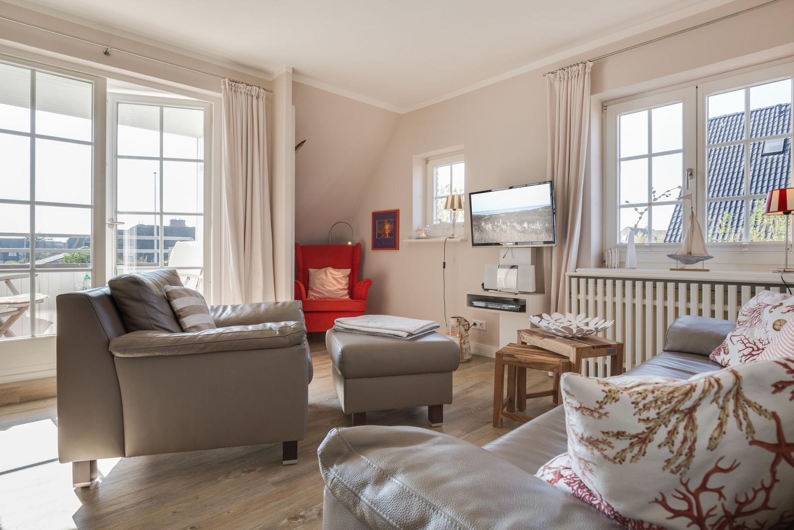 Wohnzimmer mit Sofa, Sessel und TV. Große Fenster und Holzboden.