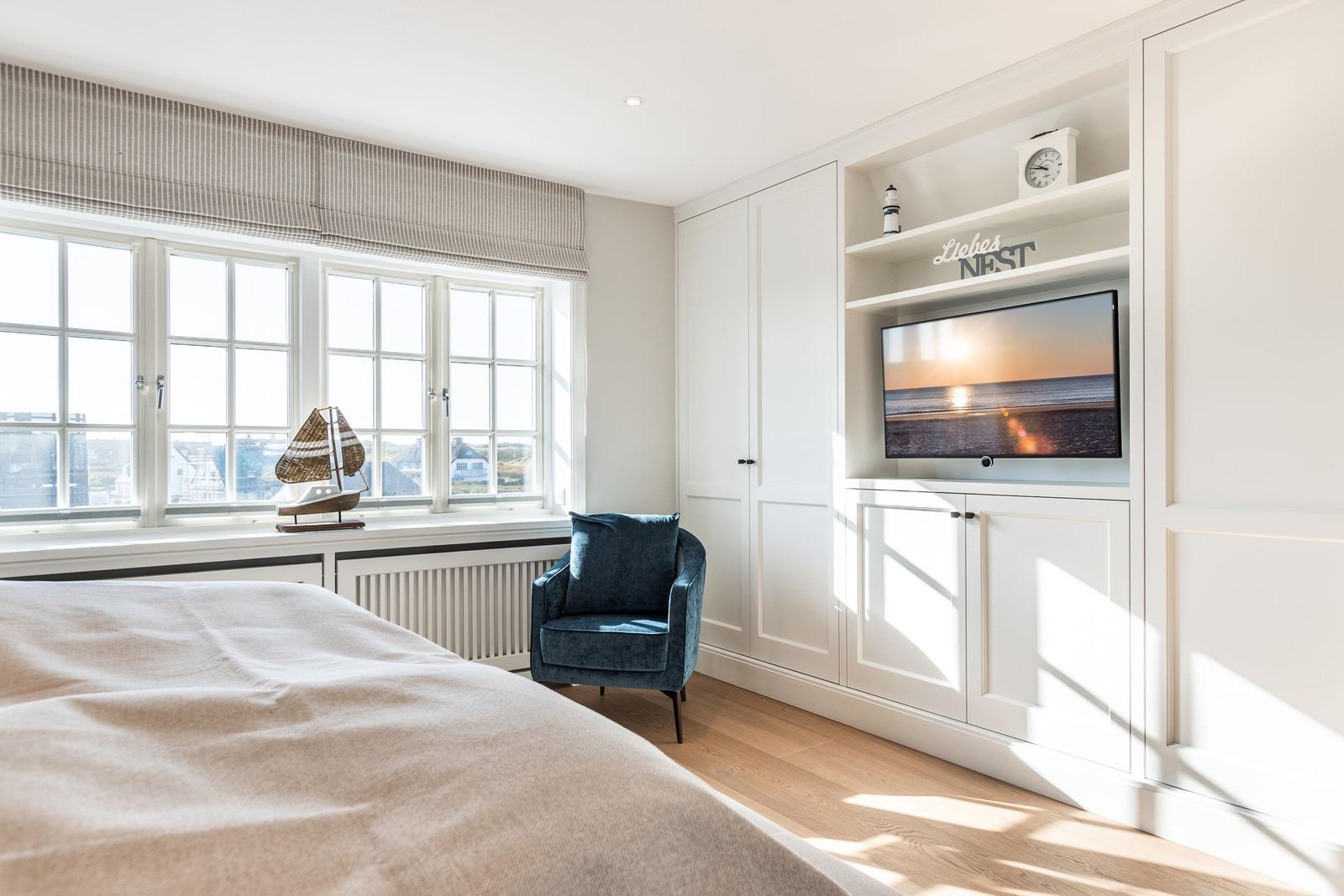 Bedroom with large window, blue armchair, and built-in TV area.