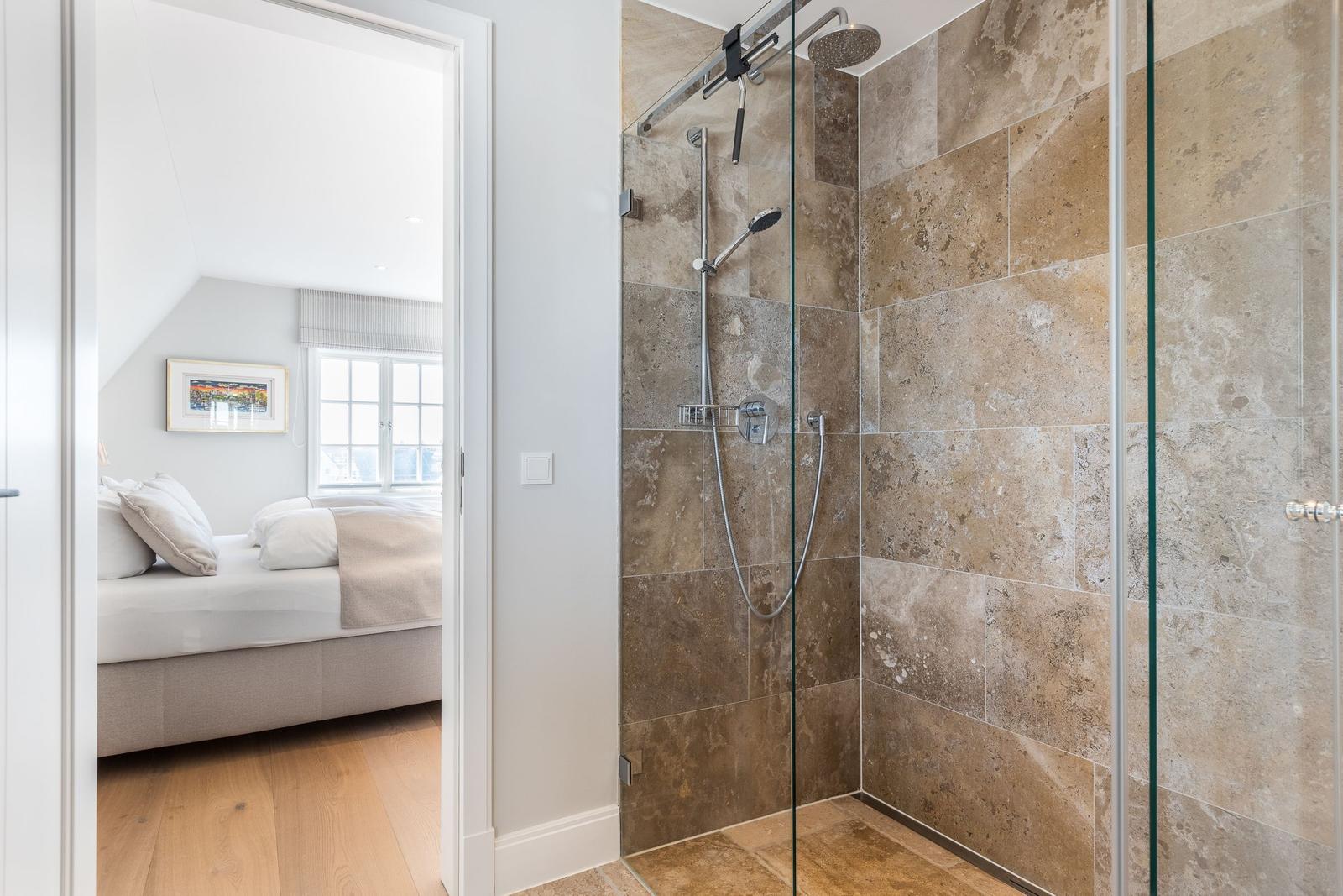 Shower with glass enclosure and stone tiles. Bedroom with bed and window visible in background.