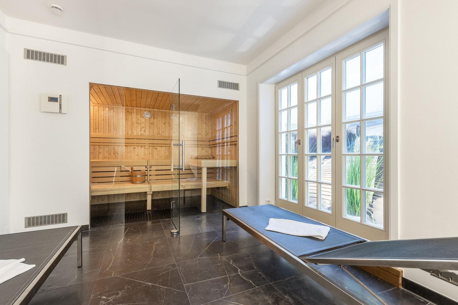 Sauna with wooden bench and glass wall. Outdoor area with windows and loungers.