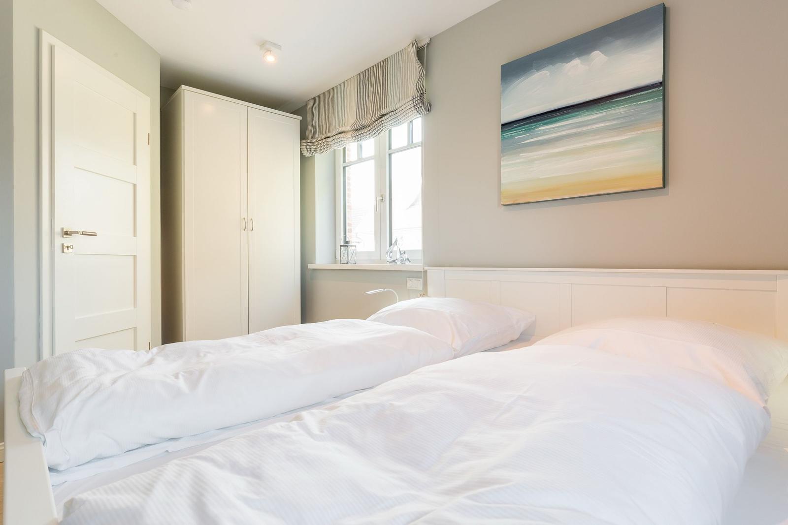Schlafzimmer mit weißem Bett, Kleiderschrank und Fenster. Wandbild zeigt Meereslandschaft.