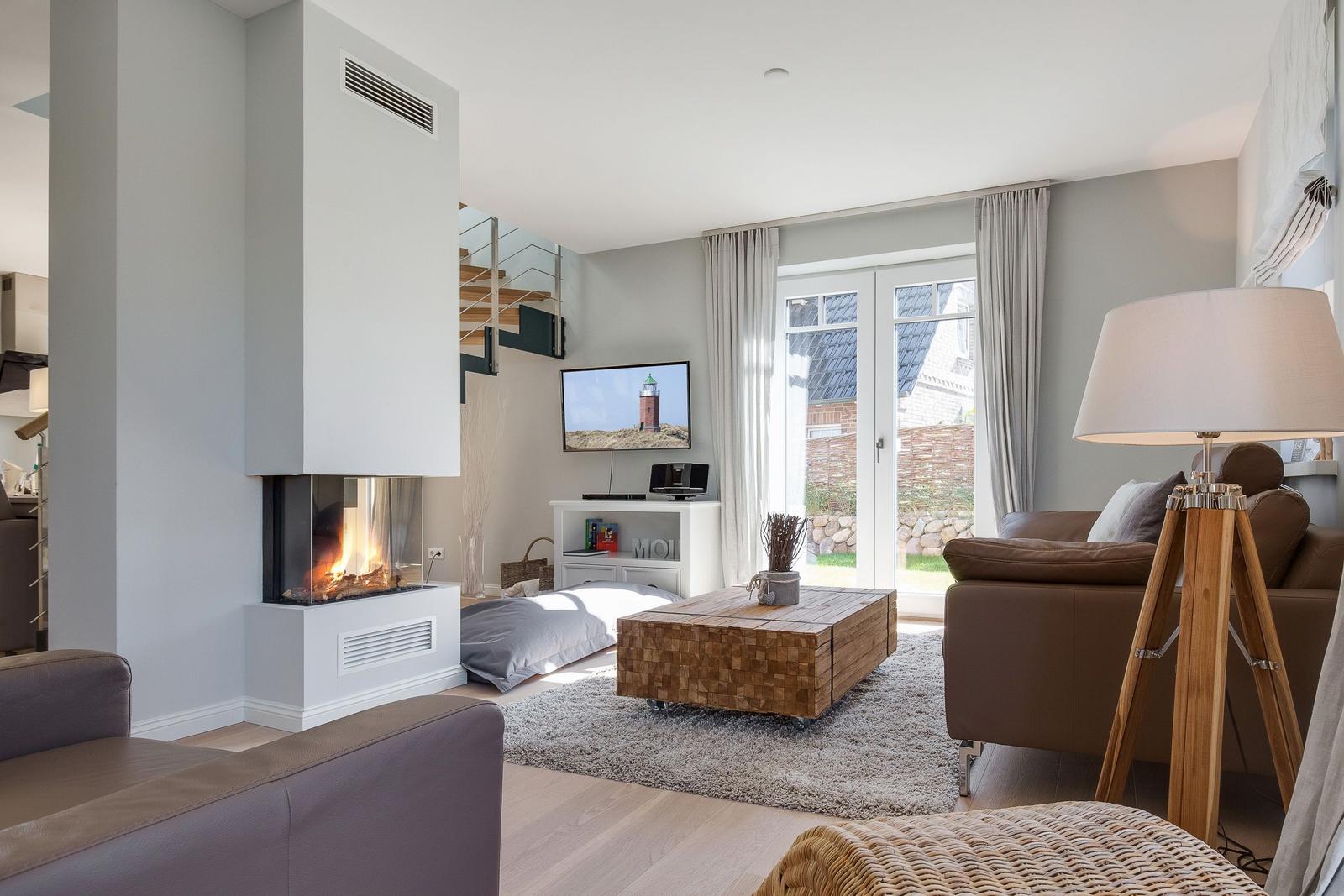 Wohnzimmer mit Kamin, Sofa und TV. Durchgang zur Küche. Terrasse mit Blick aufs Haus.