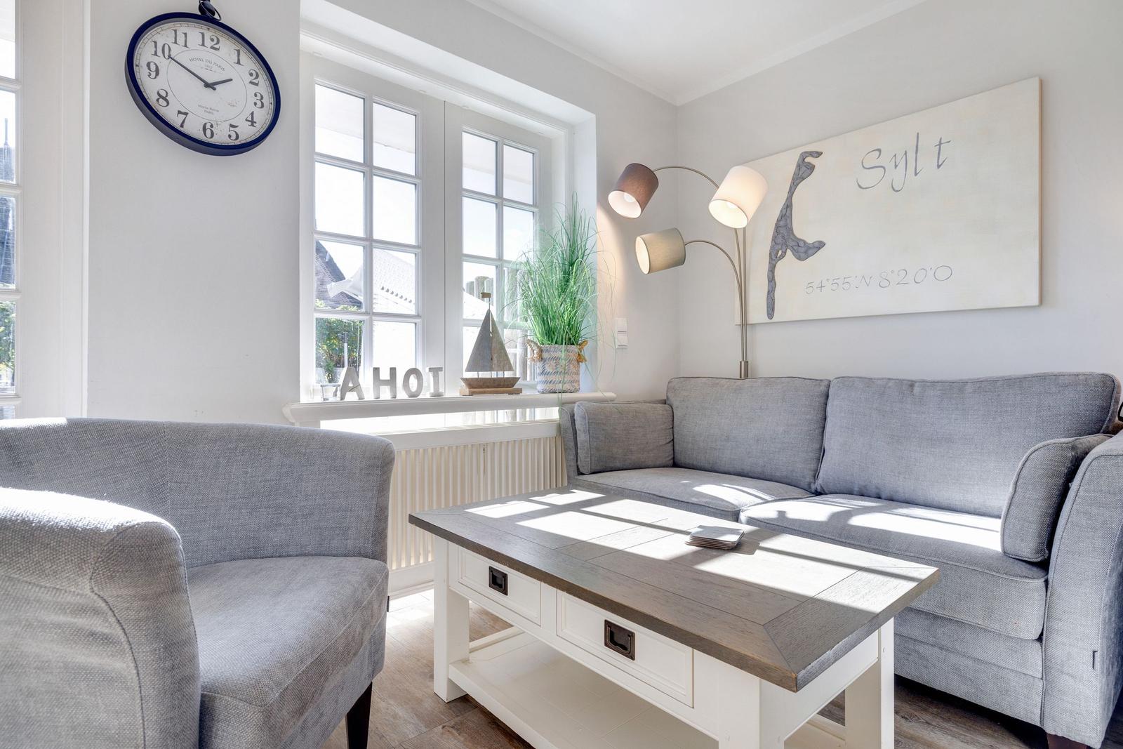 Wohnzimmer mit grauem Sofa, Tisch und Wanduhr. Fenster mit Pflanze und Wandbild.