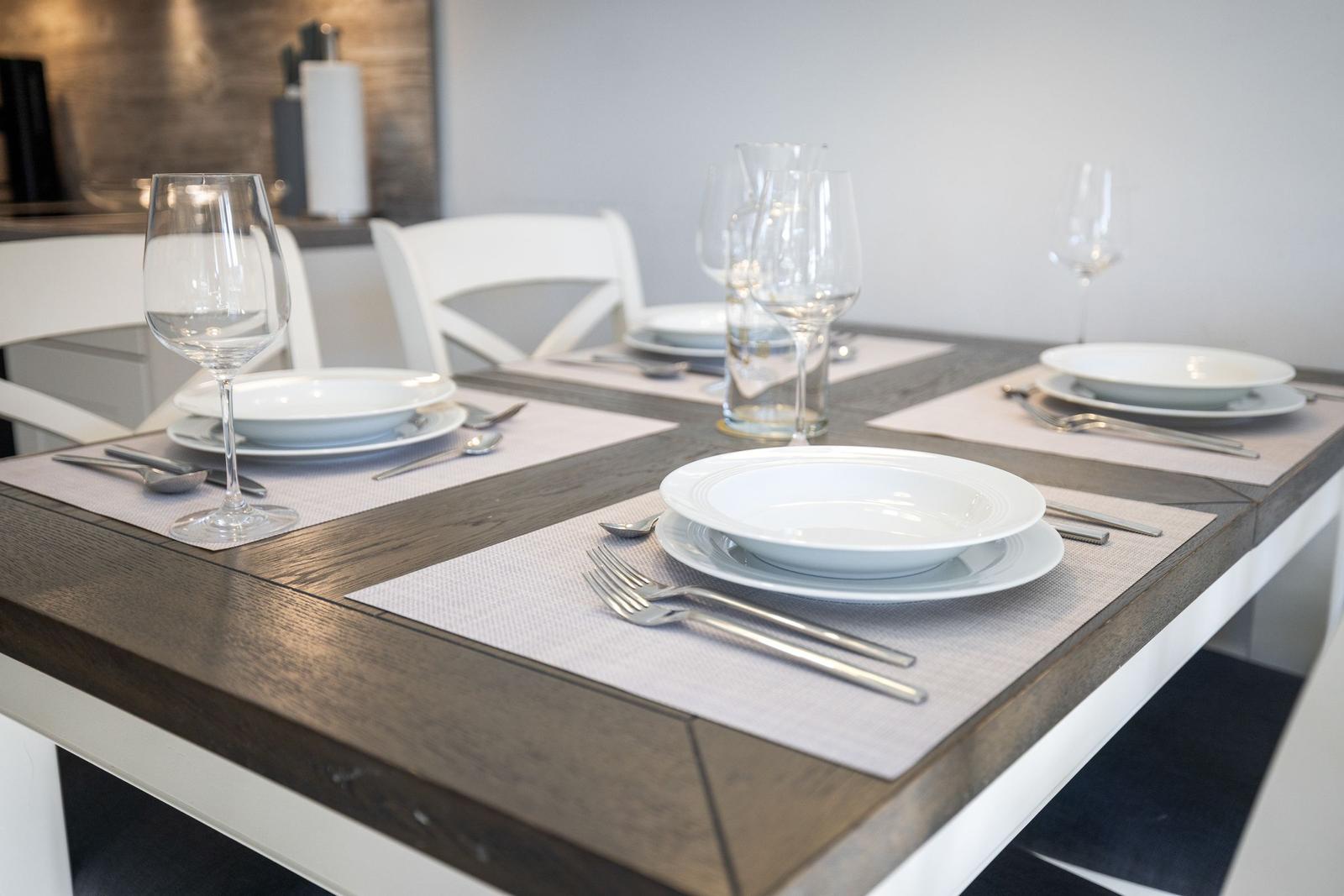 Esszimmertisch mit Platz für vier Personen, weißer Tisch, Holzoptik, Teller, Gläser und Besteck.
