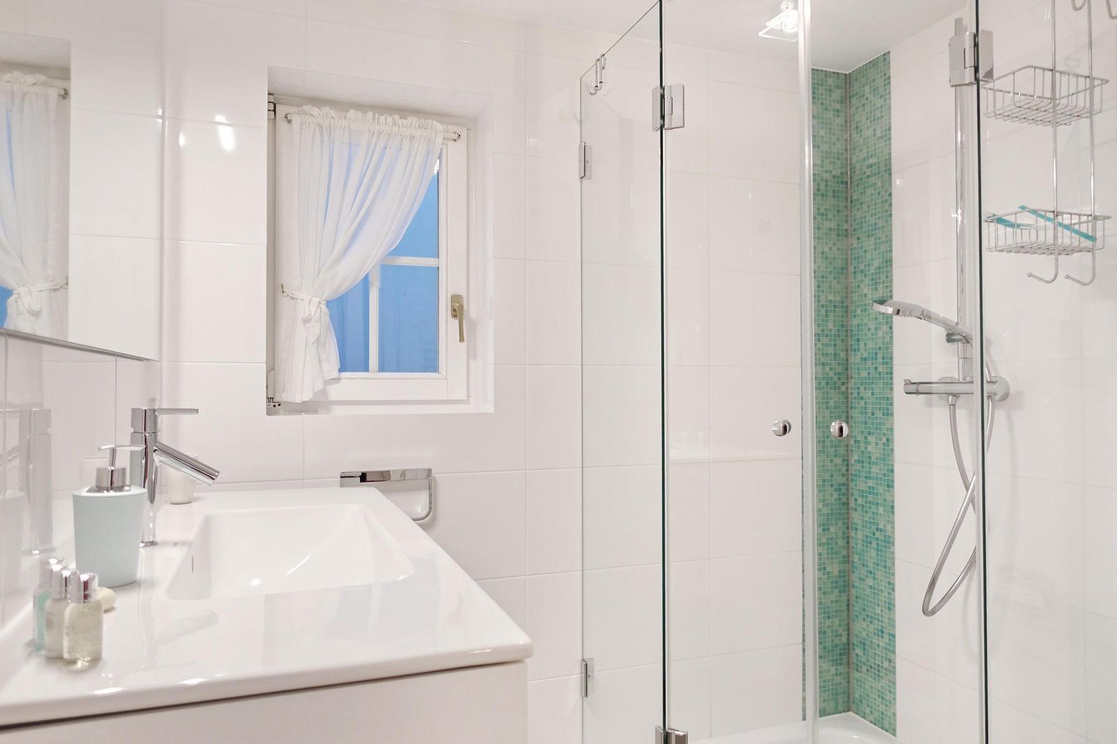 Badezimmer mit Dusche, Waschbecken und Fenster mit weißen Vorhängen.