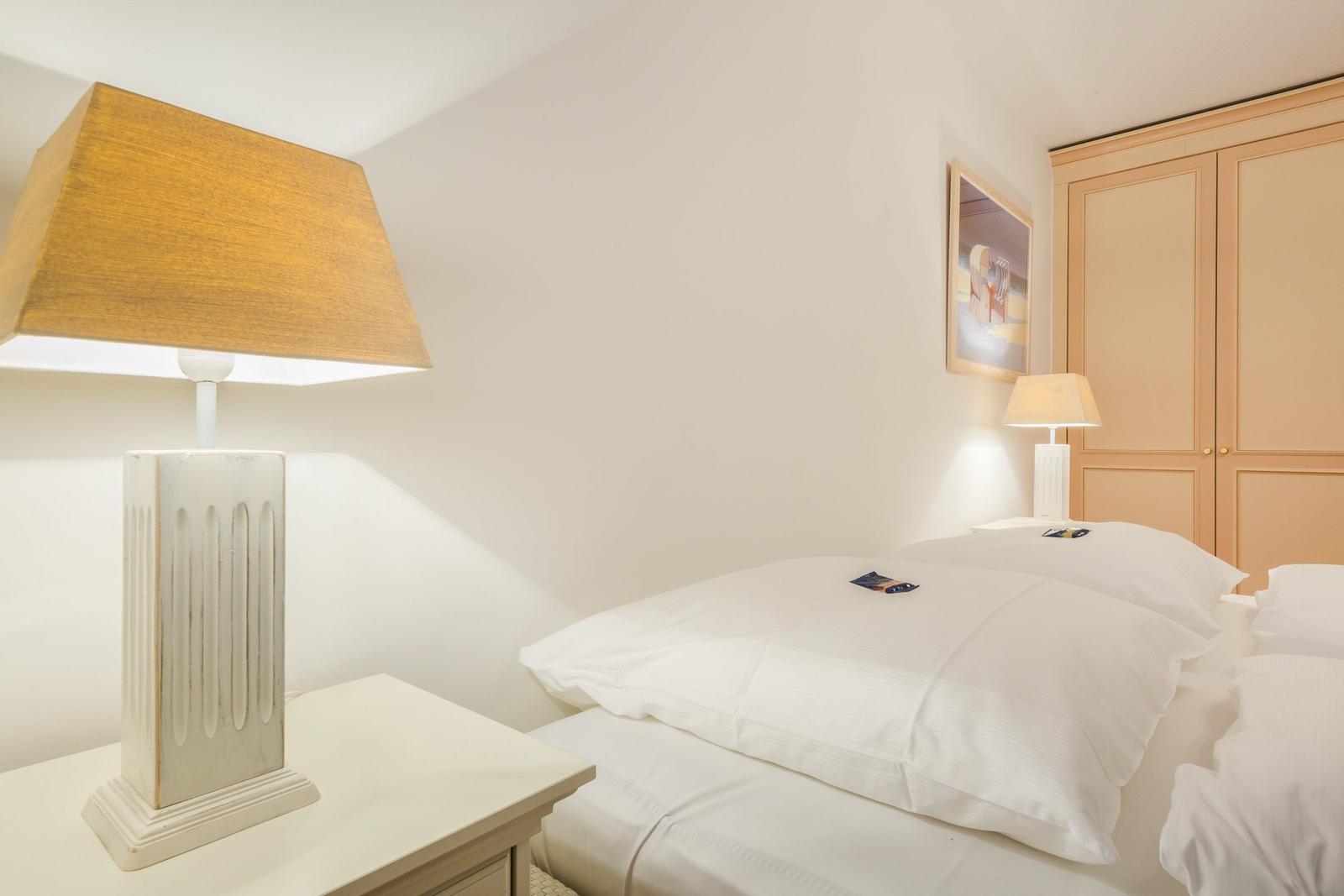 Schlafzimmer mit weißem Bett, Nachttisch und Lampe. Hinter dem Bett steht ein Schrank.