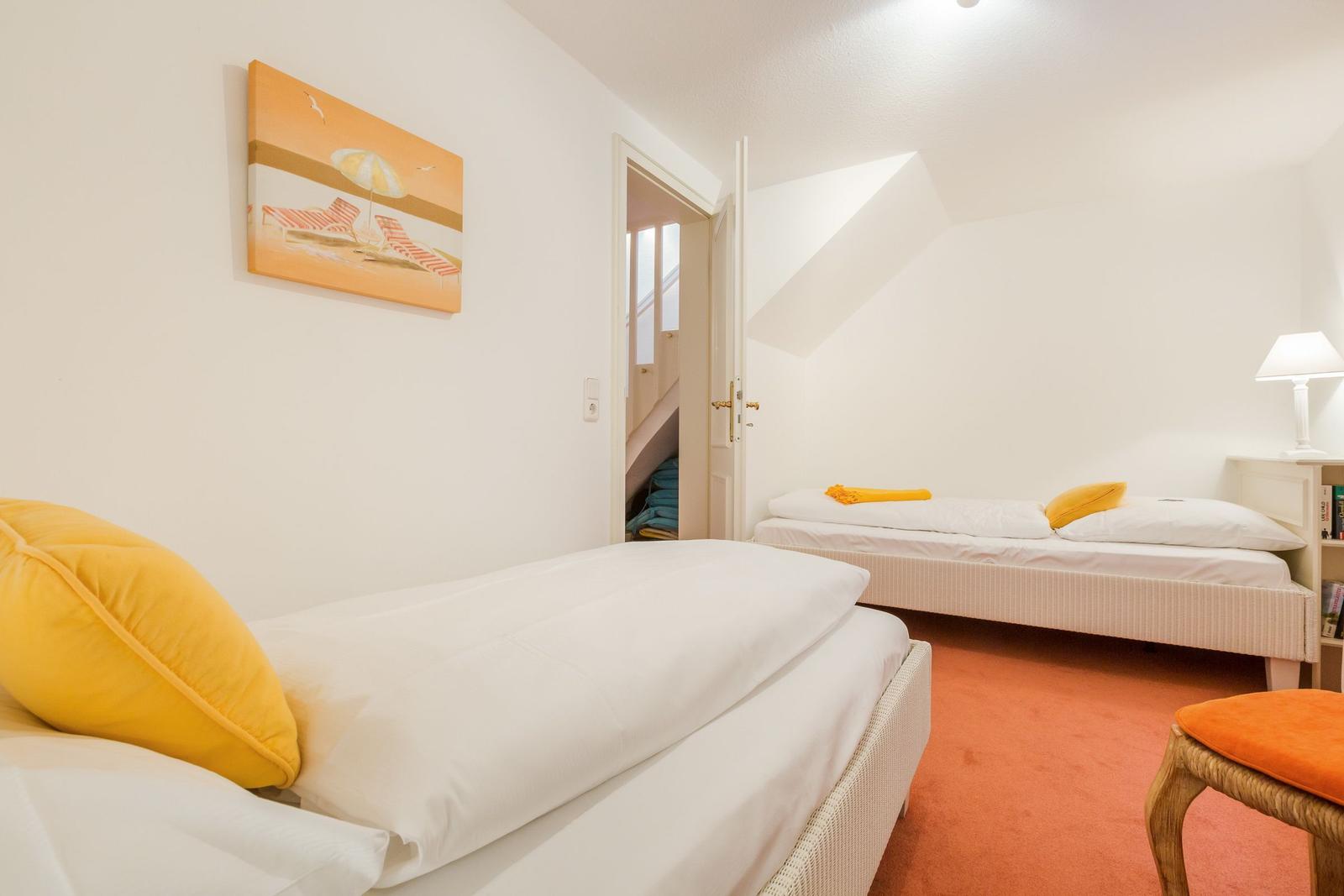Zwei Betten mit weißen Laken und gelben Kissen. Orange Teppich und Wandbild mit Strandmotiv.